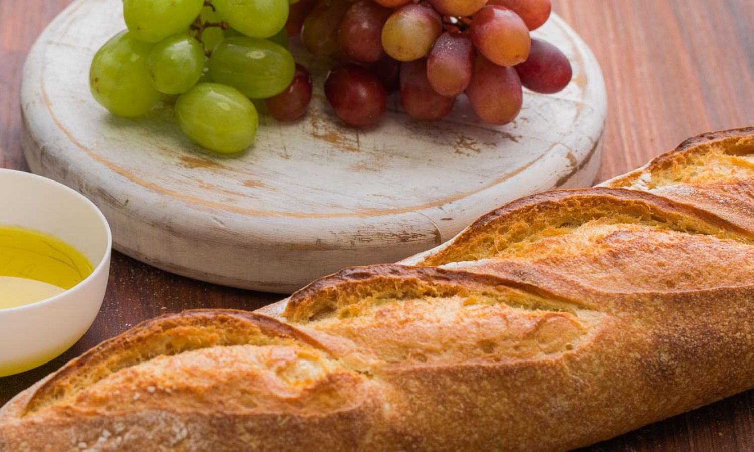 A loaf of bread, white and dark grapes and pure olive oil in a bowl. Bertolli extra virgin olive oil. Best olive oil brands.