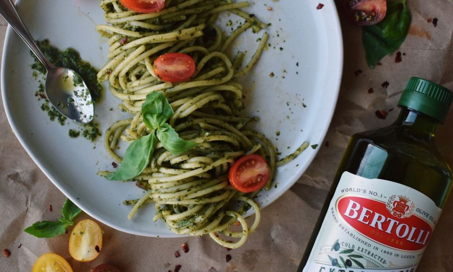 Cooked pasta on a plate alongside Bertolli Olive Oil bottle and tomatoes. Bertolli Extra Virgin Olive Oil.